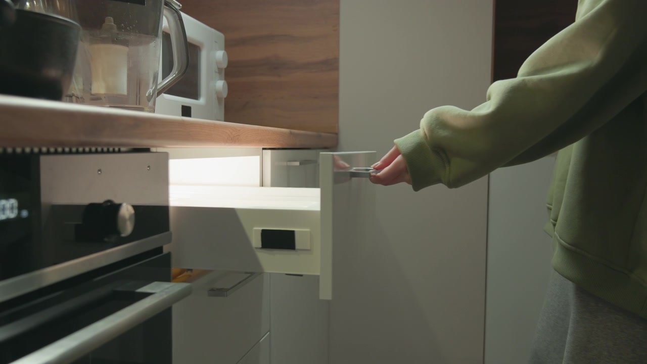 Vista ravvicinata di una persona che indossa un maglione verde che si allunga per estrarre un cassetto inferiore accanto a un forno in acciaio inossidabile, con la mano sulla maniglia, un piano di lavoro in legno, un mobile bianco e un interno di cucina moderno e ordinato.