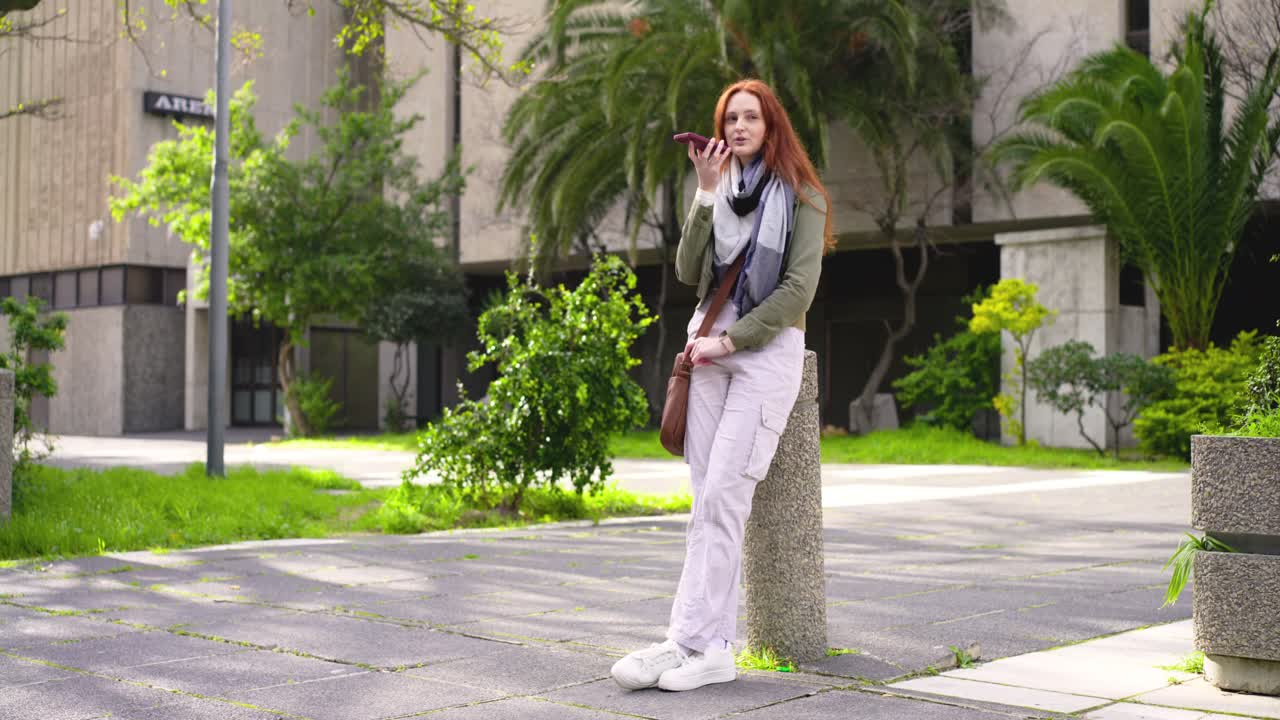 Donna dai capelli rossi appoggiata a un dissuasore nella piazza del campus, che avvia una nota vocale sul telefono mentre un passante attraversa.