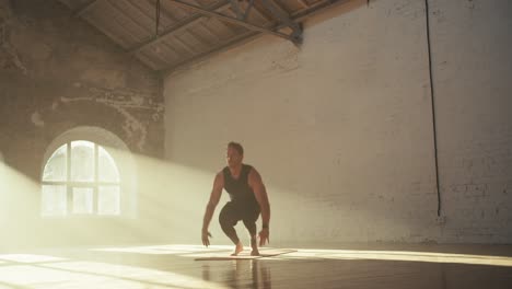 a guy in a black sports summer uniform does a burpee exercise on the red mat in the sunny brick hall