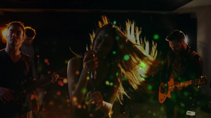 Una cantante si scuote i capelli creando particelle di luce colorate mentre la band prova musica.