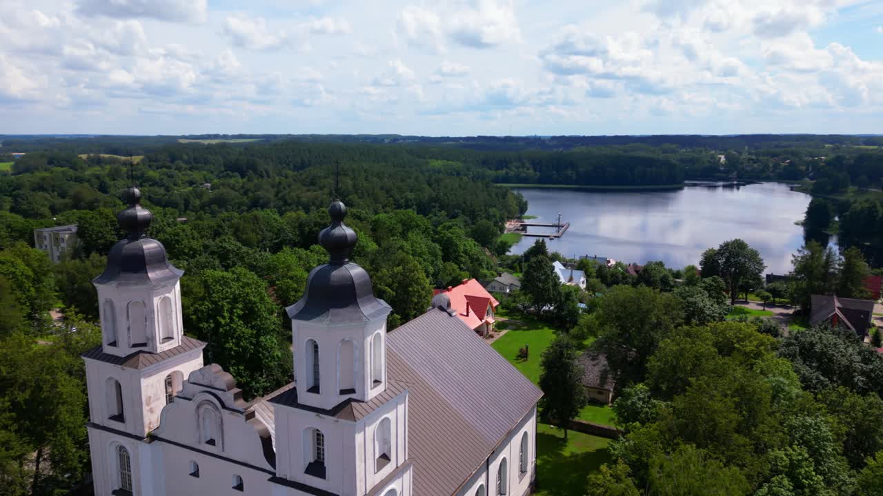 Front view of a white baroque church with black domes, framed by green trees and overlooking a calm lake. Shot at Zarasai, Lithuania (Zarasai, Lietuva)