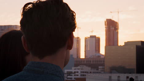 rear view of couple on rooftop terrace looking out over city skyline at sunset