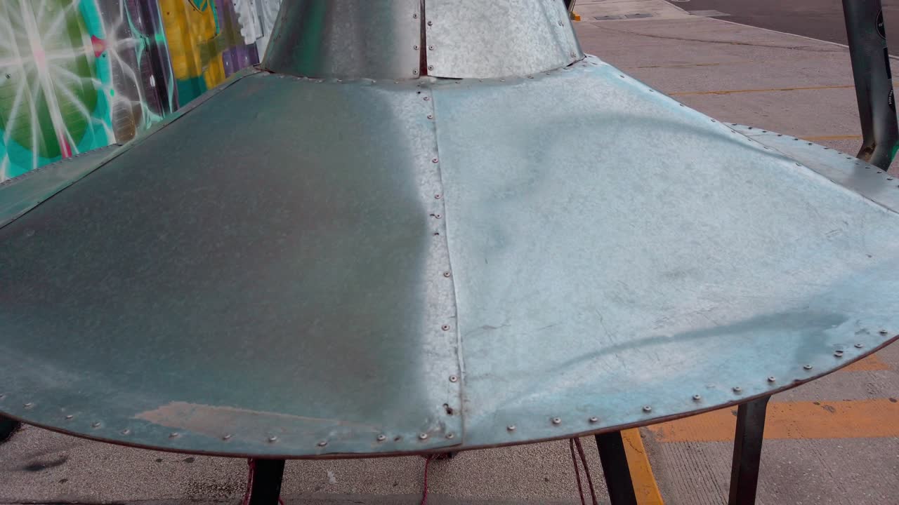 Zoom auf ein silbernes UFO-Modell vor einem Geschäft mit Graffiti-Kunst und Requisiten zum Thema Außerirdische, bei Tageslicht.