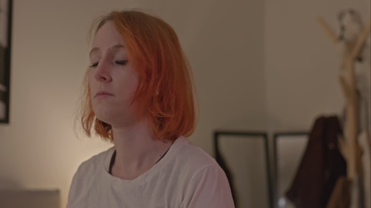 женщина с оранжевыми волосами медитирует в спальне, когда камера следит за ней.