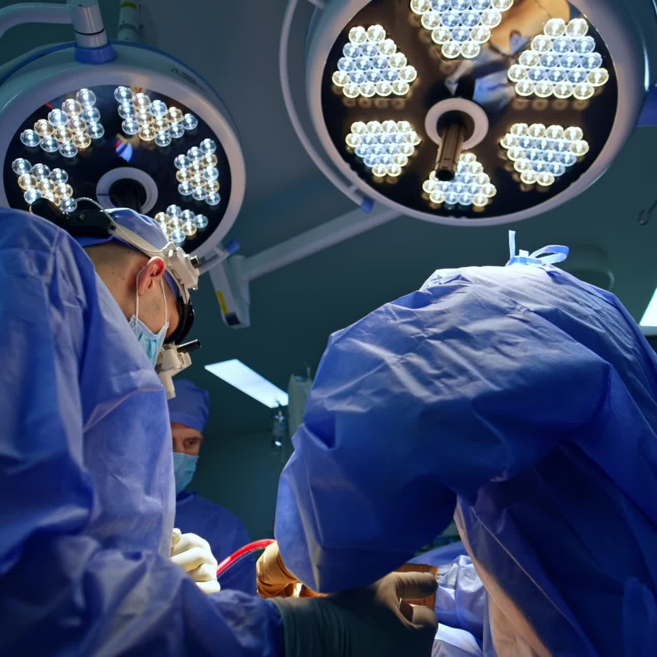 Rear view of two neurosurgeons cooperating at surgery. Male assistant passes the tool to the doctor. Round lamps at backdrop. Low angle view