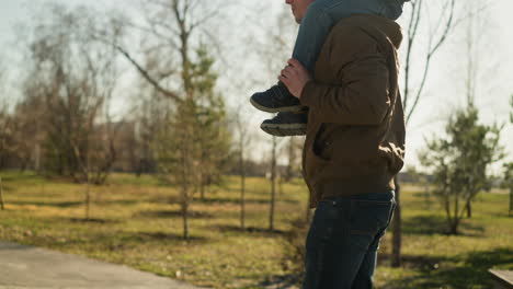 a close-up of a father walking with his son on his shoulders, focused on where they are headed, as they walk on a paved path near an open field, the father wears jeans and black sneakers