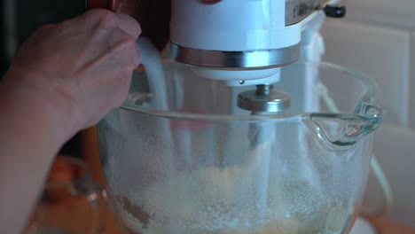 woman adds sugar ingredient to a moving electric stand mixer with flour in glass bowl