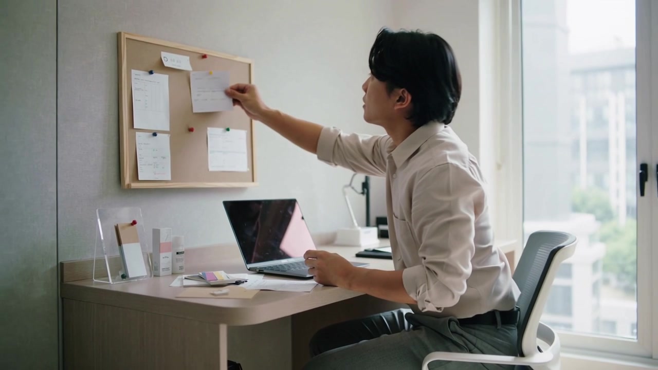 Reading small card, man wearing light shirt pinning note to corkboard at desk, organizing workspace