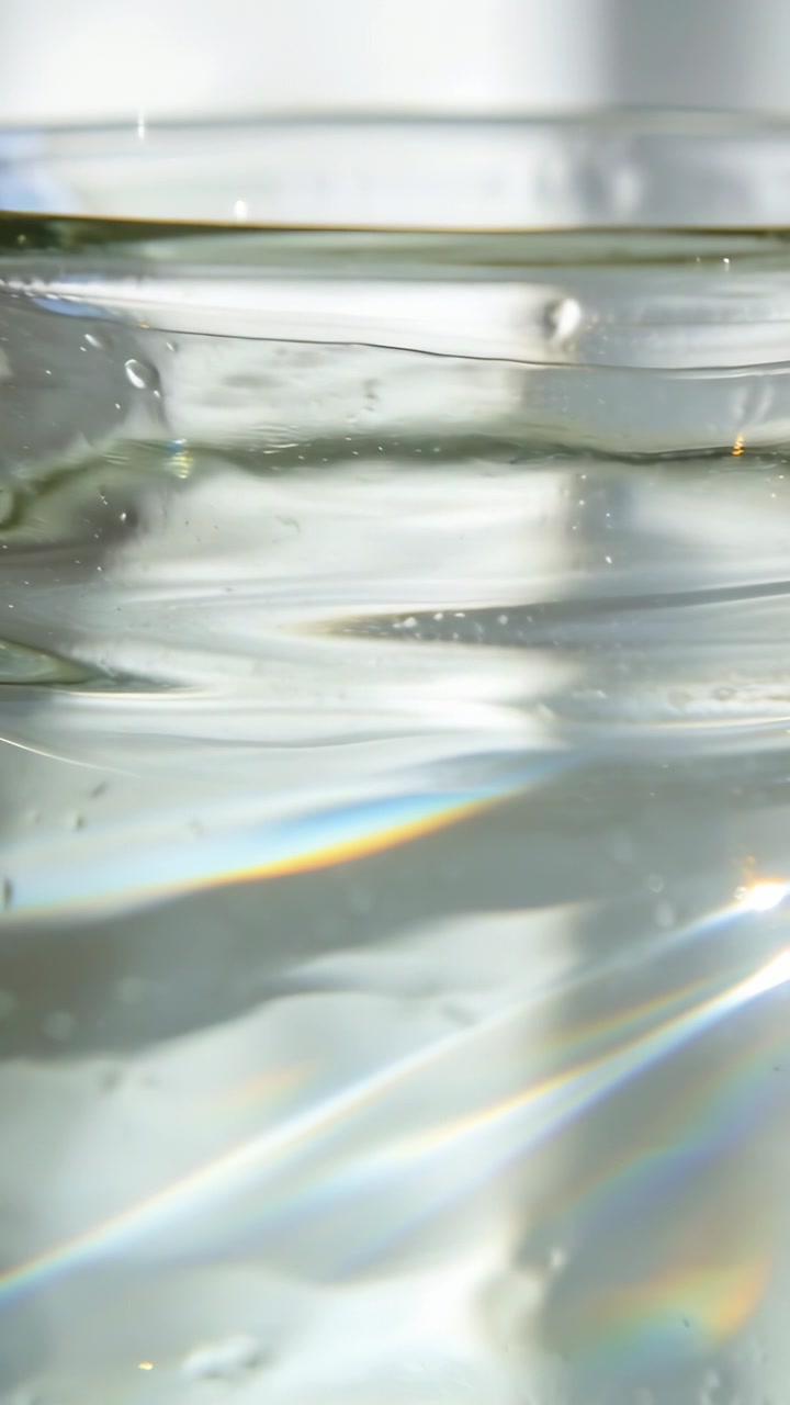 Vertical video: Rippling water in glass on desk by window as light moving, bubbles and prism band
