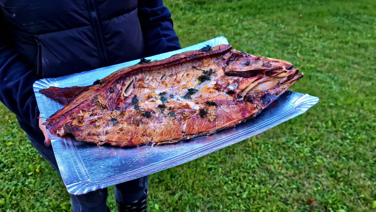 An unrecognizable woman in a black jacket carries a tray of cooked carp, close up