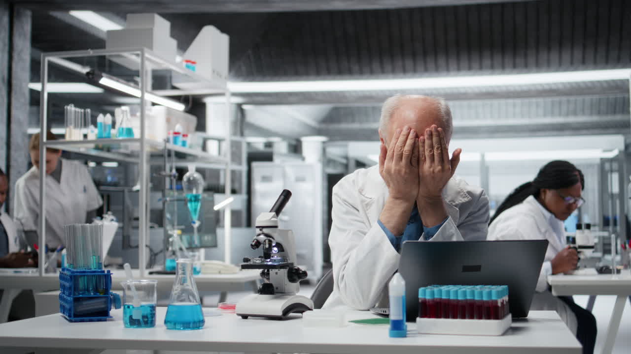 Ricercatore anziano di nanotecnologie di laboratorio che si sente sopraffatto, lavorando al laptop
