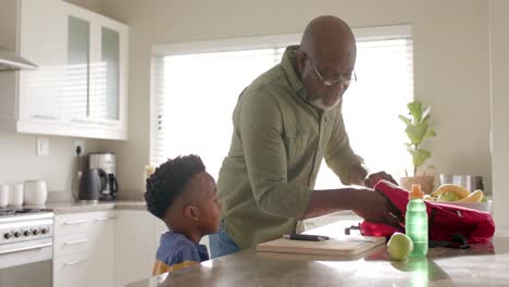 Abuelo-Afroamericano-Preparando-Almuerzo-Para-Llevar-Con-Su-Nieto-En-La-Cocina-Antes-De-La-Escuela,-Cámara-Lenta