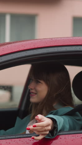 young woman drives car in rainy weather. smiling female driver enjoys comfortable trip in city. pleasant driving with open window on blurred background side view