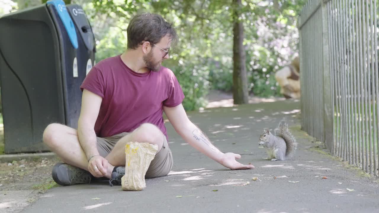 Uomo seduto su un sentiero cerca di far prendere cibo dalla sua mano a uno scoiattolo