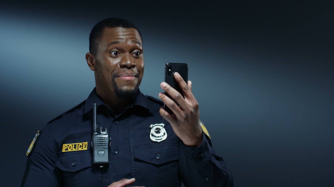 African American young cheerful handsome policeman in uniform and in walkie-talkie smiling and having videochat on the smartphone via web cam on the dark wall background.