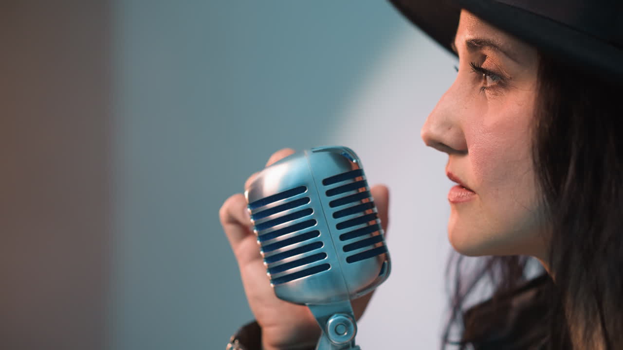 Close side view of a singer wearing a black hat and holding a vintage microphone.,singer's facial profile and expression are highlighted against a white background as the camera zooms out