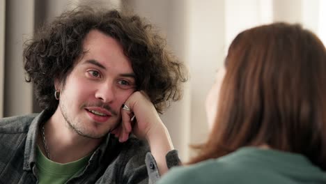 over the shoulder, a happy brunette guy with curly hair and a mustache props his head with his hand and talks to a brunette girl at home on the sofa