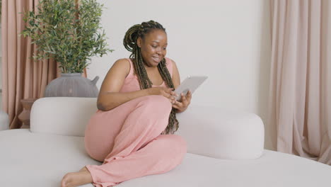 woman with braids sitting on a big sofa while using a tablet