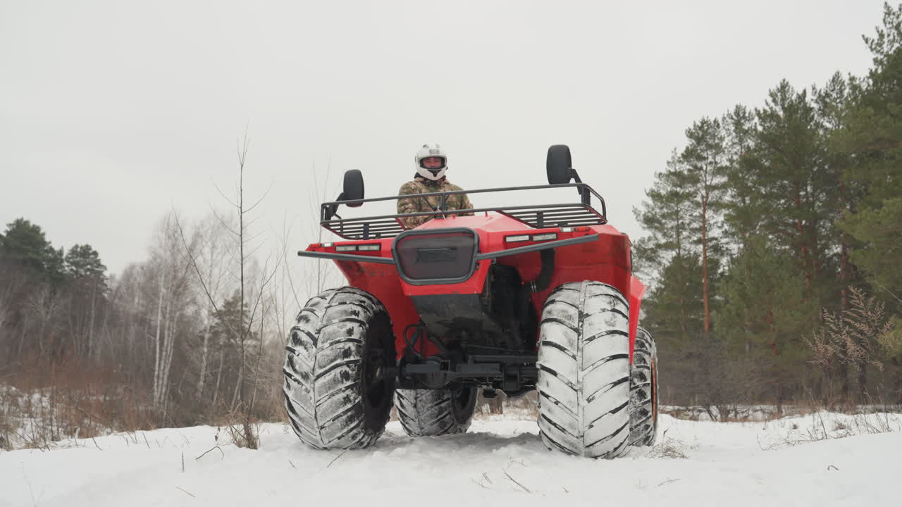 ATV rosso che sfreccia su un sentiero innevato, un pilota solitario con casco si fa strada nella neve battuta, pneumatici con battistrada profondo sollevano fiocchi di neve, ai margini della pineta, angolazione frontale bassa, cielo coperto, atmosfera grintosa e piena di energia.