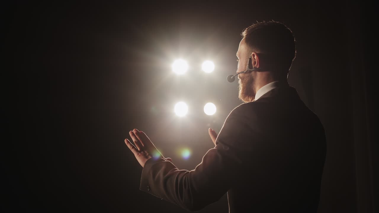 Il relatore tiene la conferenza. È in piedi sul palco e parla al microfono. Sta gesticolando. Il riflettore lo illumina. La telecamera gli sta ruotando intorno. 4K
