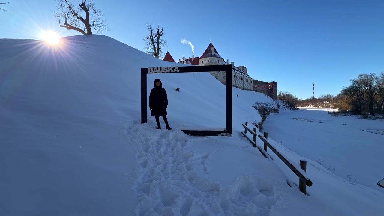 Henkilö poseeraa valokuvakehysveistoksessa Bauskan linnassa Latviassa talvisessa lumipeitteisessä maisemassa.