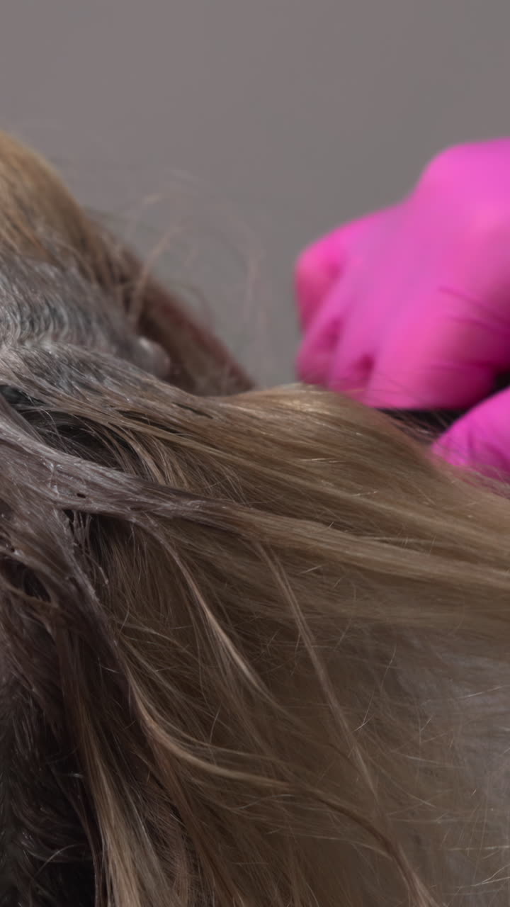 Primo piano stilista che applica il colore alle radici della scriminatura di una cliente bionda caucasica, piccole pennellate ripetute. Media