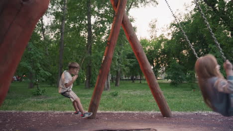 children swinging freely in leafy playground at dusk with blurred forest background capturing dynamic motion joyful laughter summer fun outdoors under warm ambient light with wooden swing frame