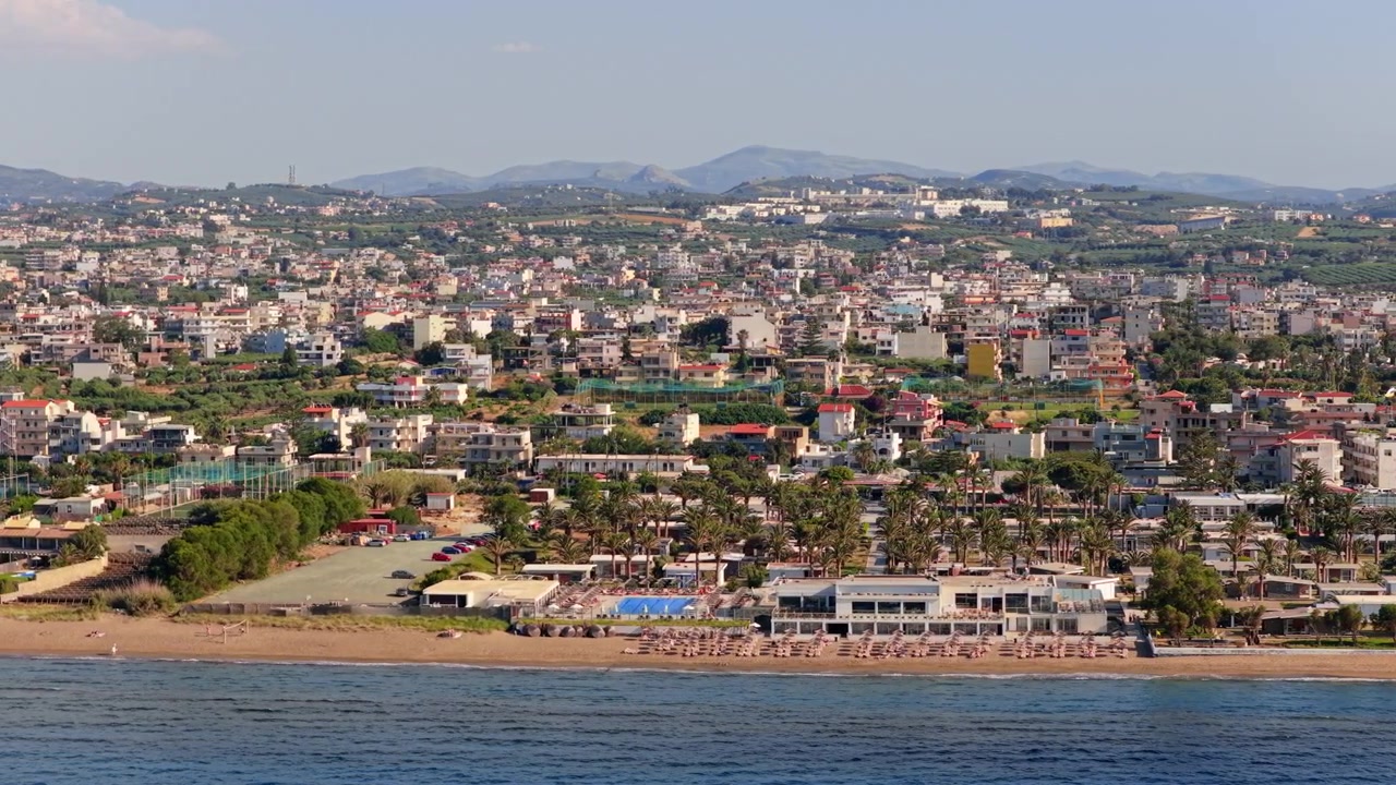 Vista aerea con drone di una spiaggia di resort con ombrelloni di paglia e hotel fronte mare sotto una città e colline verdi, Ammoudara, Grecia