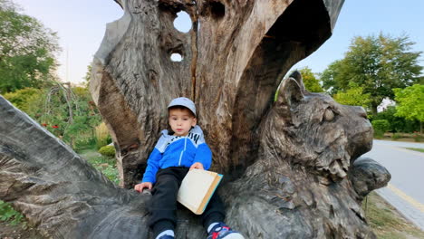 adorable baby boy sitting on the wooden cat sculpture. cute toddler with a book in the park.