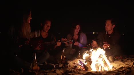 multiracial group of young boys and girls sitting by the bonfire late at night and singing songs and playing guitar. cheerful