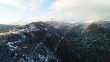 Aerial-view-of-TV-tower-on-mountain-top-surrounded-with-forest-covered-in-snow