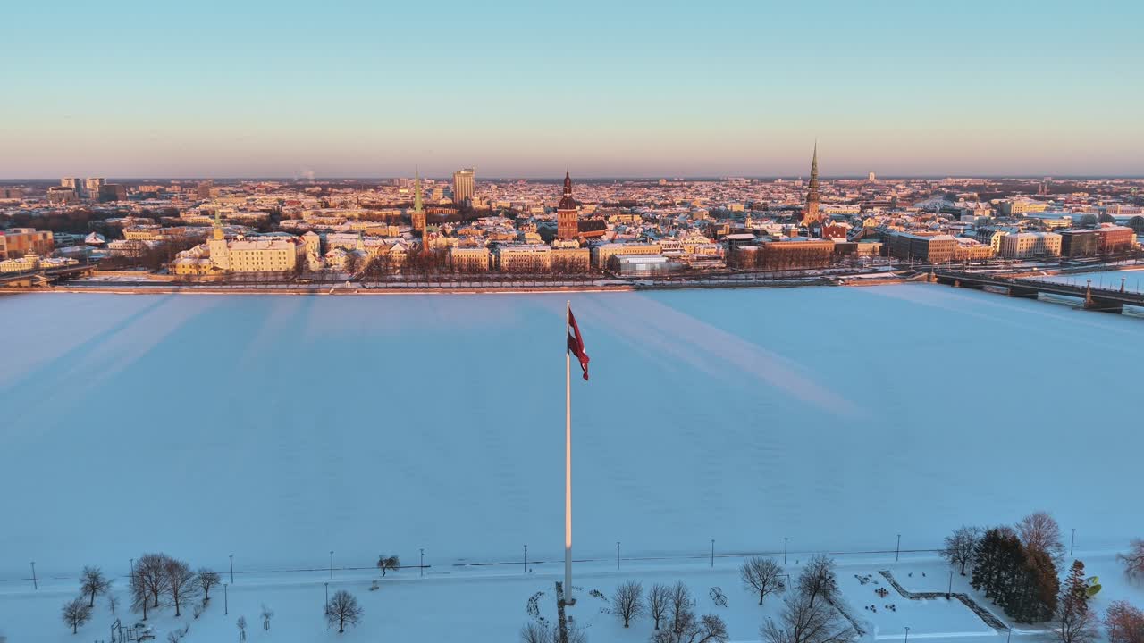 Bandiera lettone che sventola sopra un fiume ghiacciato con lo skyline di Riga coperto di neve.