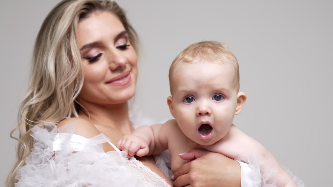 Adorable cute Caucasian infant baby opening mouth. Little child in mom's hands. Mother and son studio footage. White backdrop.