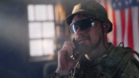 male soldier in combat gear talking on tactical phone with american flag in background