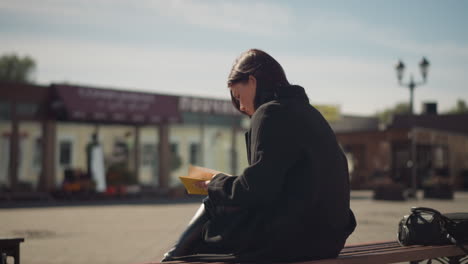 woman in black coat sitting outdoors on a bench, reading a book, with yellow-edged pages visible, background features urban buildings, lamp poles, and some plants