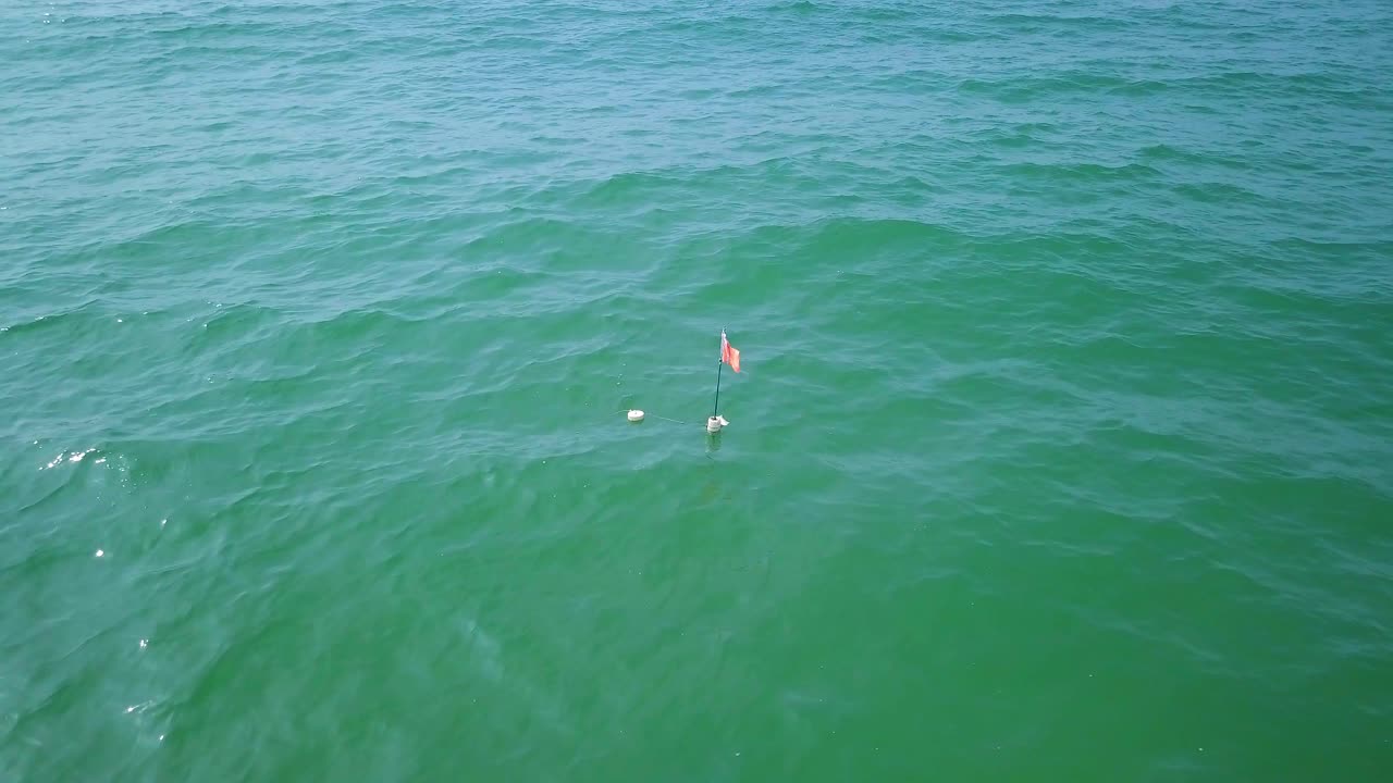 Aerial birdseye view of lush green Baltic sea in a sunny summer day, a fisherman's net buoy with the red flag floats on the top of the water, small waves moving slow