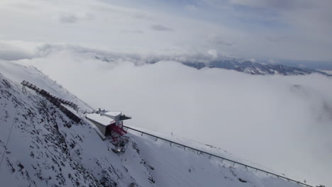 Toma-Aérea-De-Arriba-Hacia-Abajo-Del-Ascensor-En-El-Pico-De-La-Montaña-Nevada-Por-Encima-De-Las-Nubes-En-Invierno---Austria,-Europa