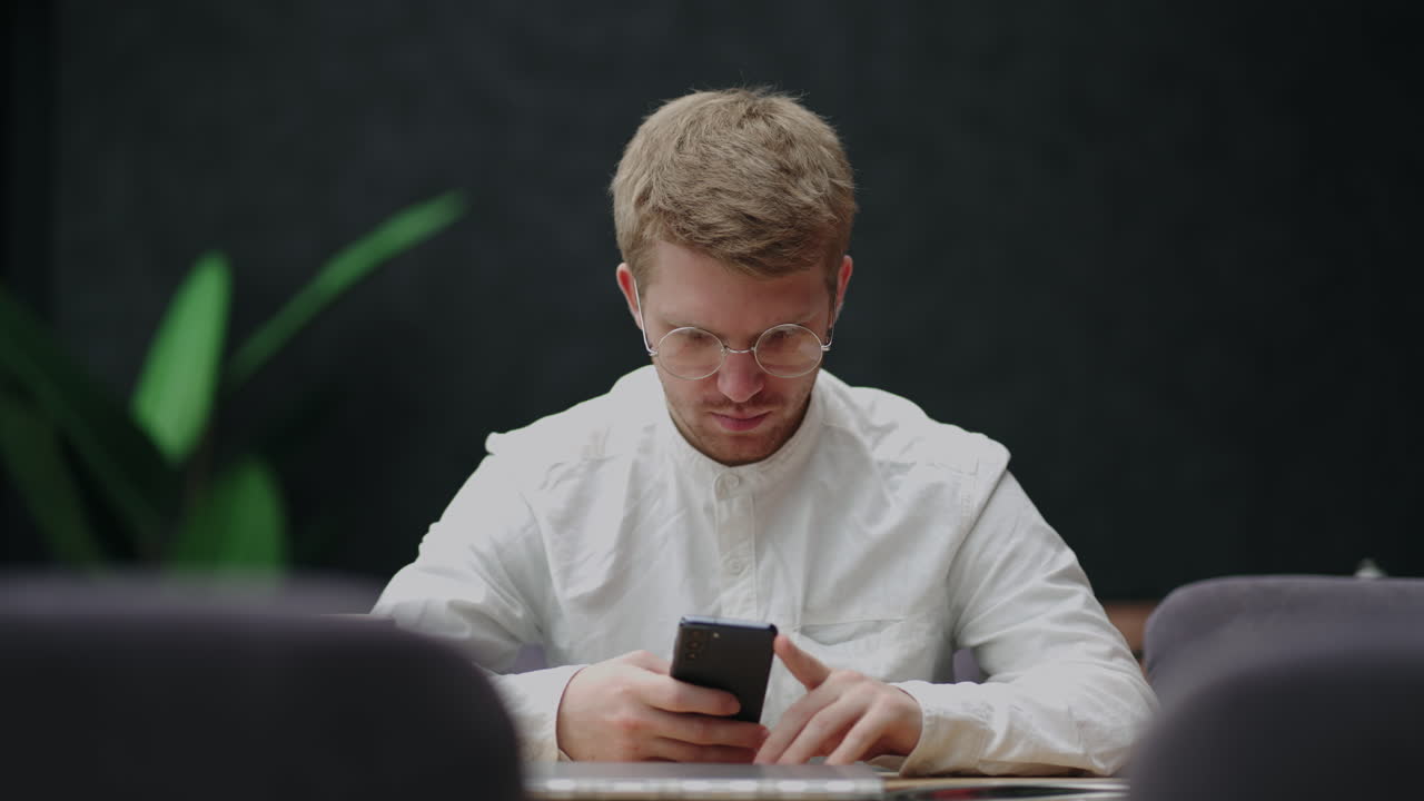 молодой человек в очках с мобильным телефоном в руках смотрит на экран и отправляет сообщения мужчина пользователь