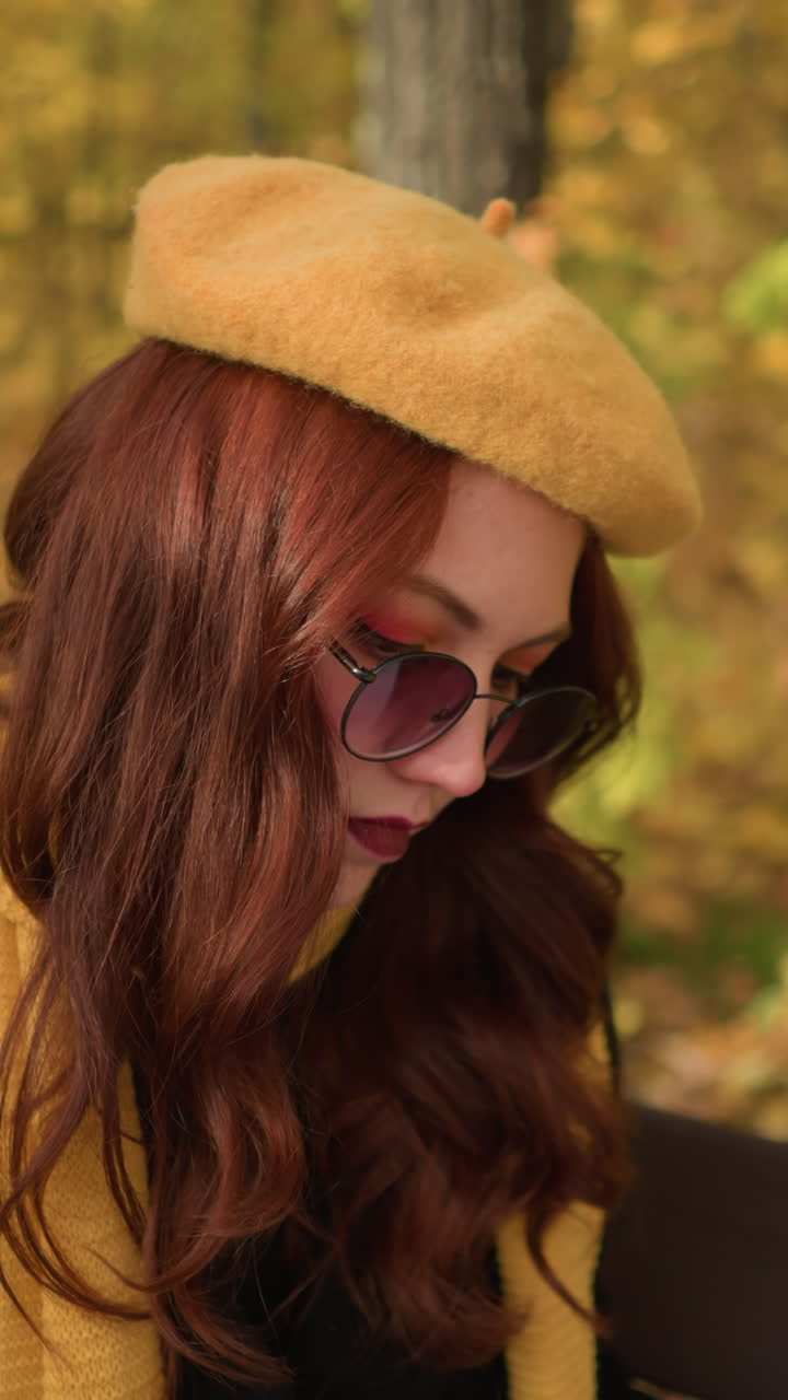 노란색 베레, 선글라스, 은 파란 머리카락을 입은 여인이 캔버스를 바라보고, 머리를 오른쪽에서 왼쪽으로 기울이고, 예술적인 생각에 빠져, 황금한 가을 잎자루로 둘러싸인 나무 벤치에 앉아 있습니다.