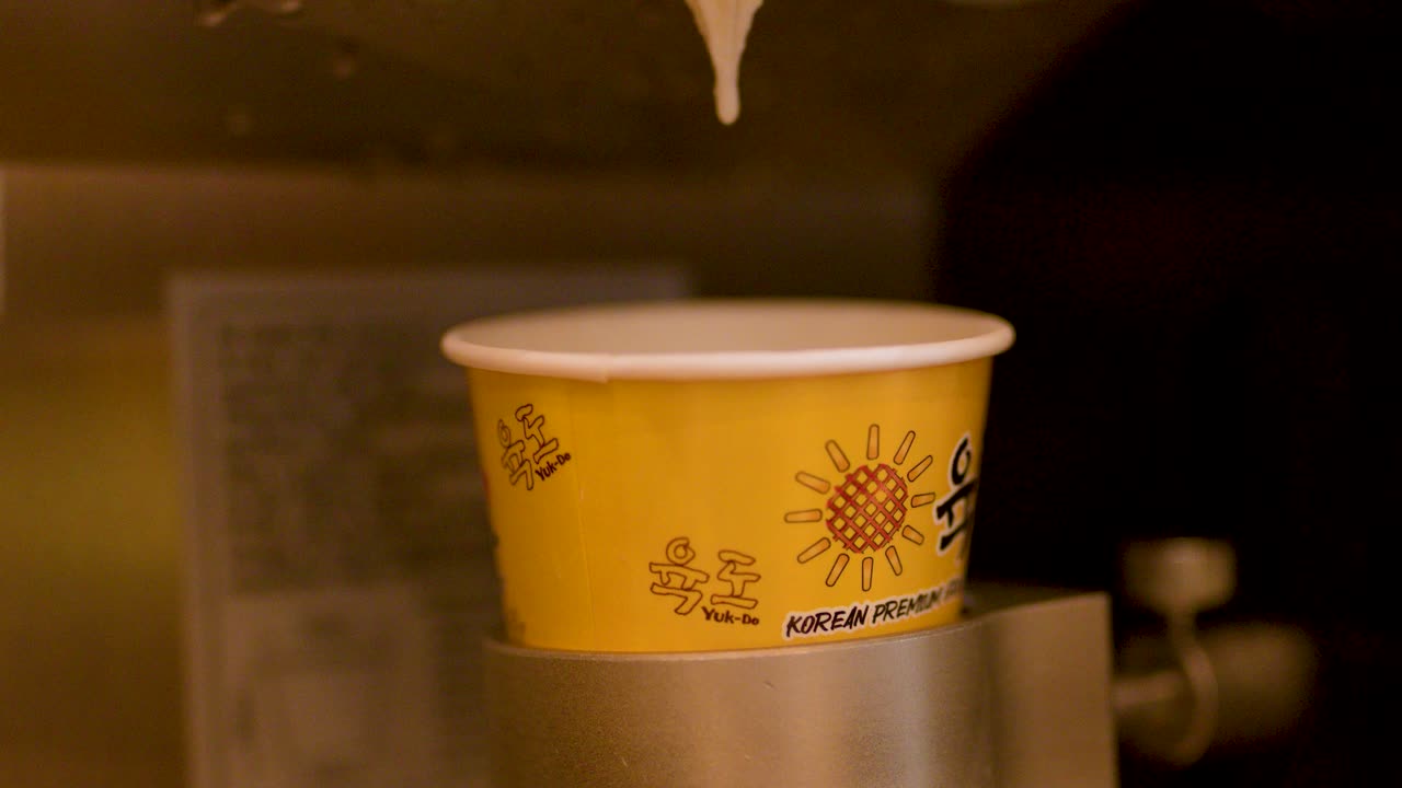 Hand positions paper cup under dispenser, machine releases dessert, warm indoor lighting, close-up view