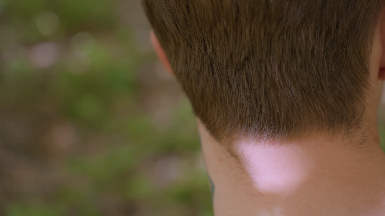Back view of white man showing ear and shaved side beard skin while untying headband, highlighting neck tattoo and detailed grooming, in natural outdoor environment with soft sunlight
