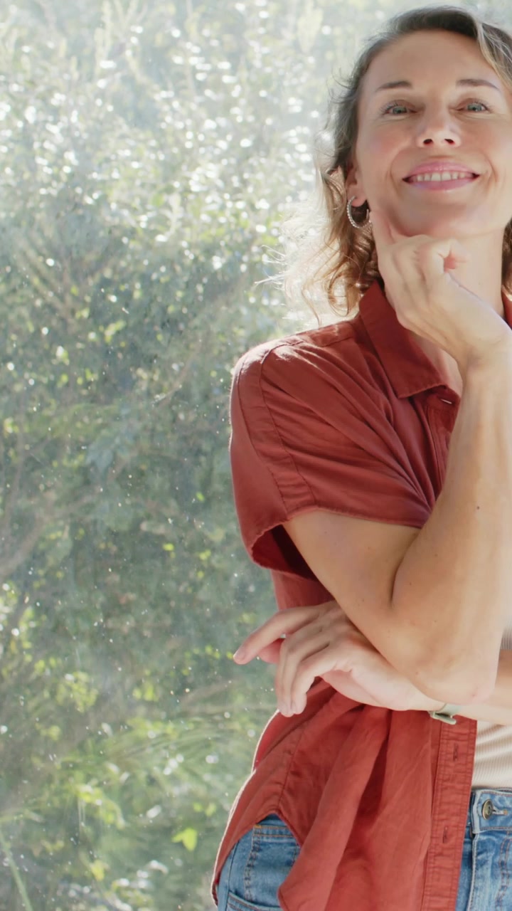 Video vertical: Mujer moviéndose con camisa color óxido, cruzando los brazos y sonriendo a la cámara junto a una ventana, con espacio para texto.