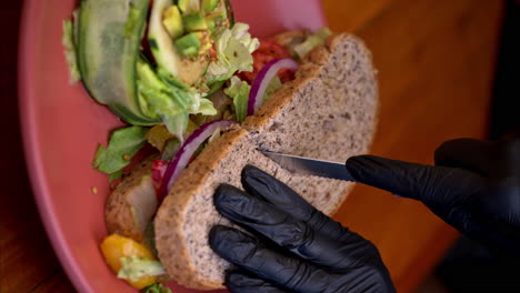 vertical slow motion of a person wearing black latex gloves cutting in half a whole wheat bread with visible onions and pickles