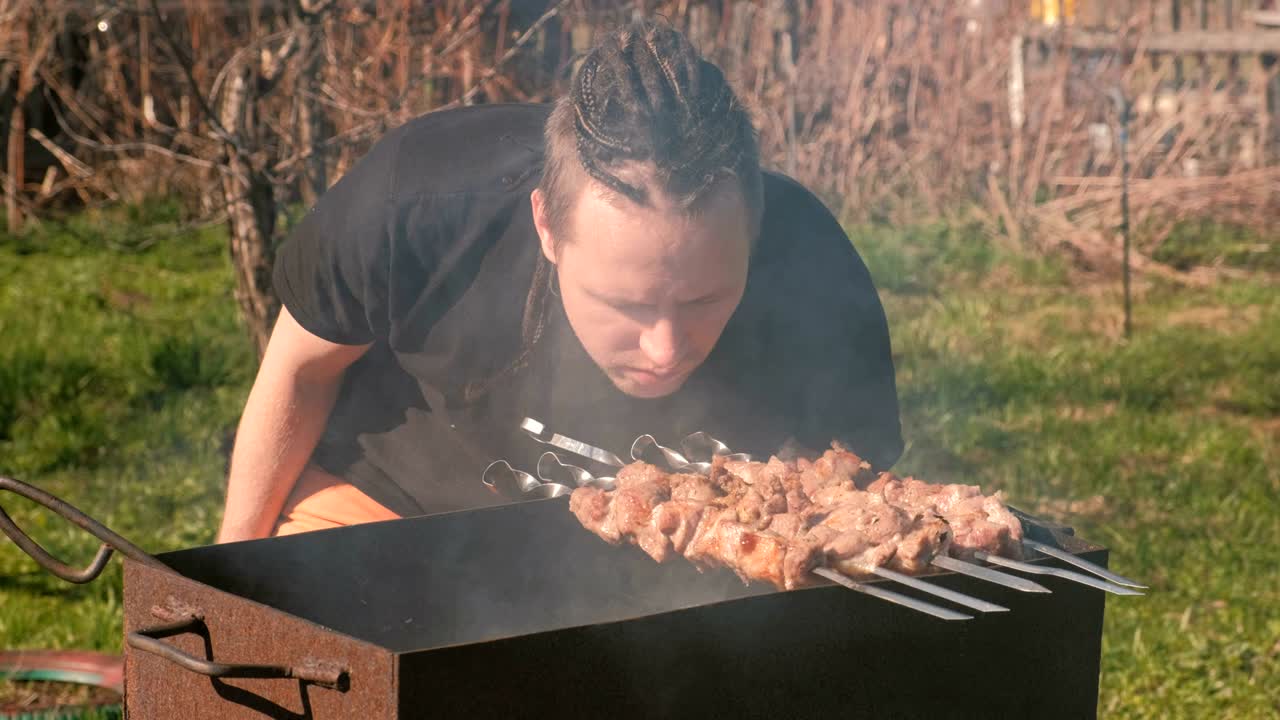 Young man with dreads on hairs cooks shashlik meat on top of charcoal grill on backyard.