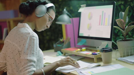 business woman working late writing notes using computer online research information data planning project deadline strategy in office at night wearing headphones