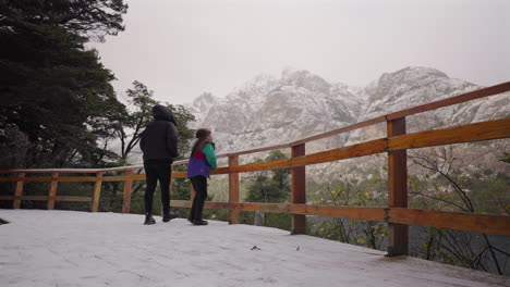 two tourists stand on a wooden observation deck in bariloche, argentina, gazing at the snow-covered peaks of the andes mountains, surrounded by alpine forest and winter atmosphere