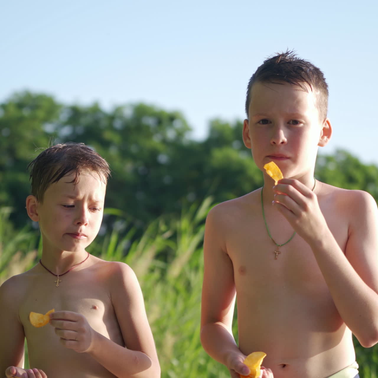Hungry boys eating on the natural green background. Two brothers with bare bodies standing outdoors and eat tasty food in summer. Happy childhood.