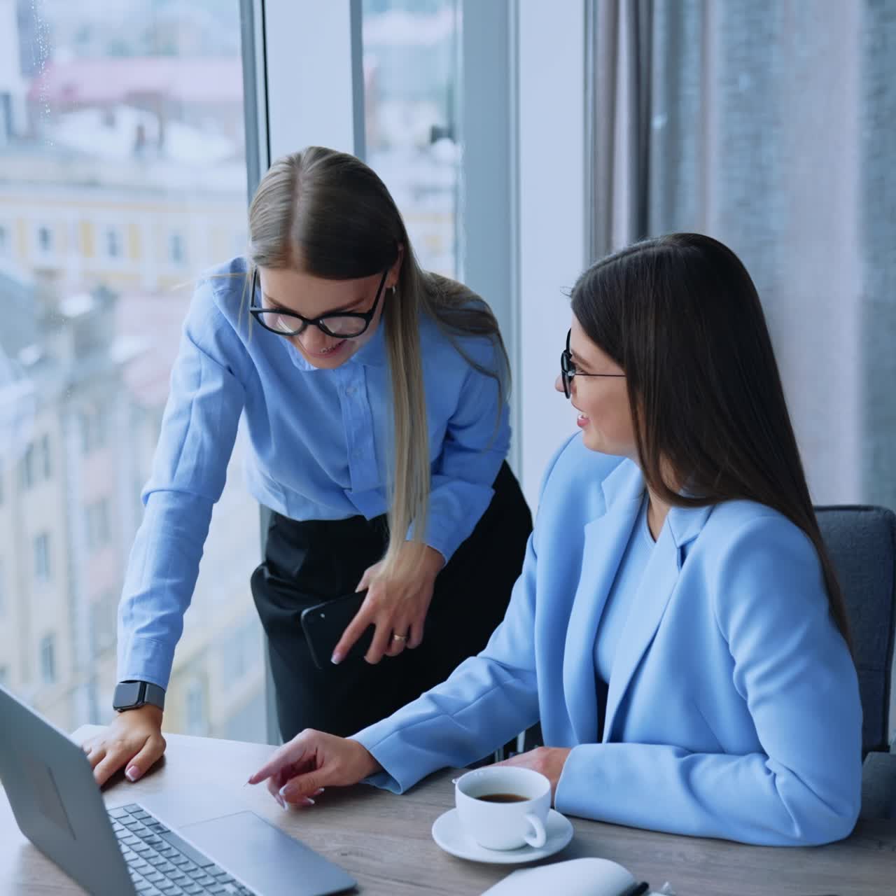 Two female colleagues collaborating to gain the best results at work. Young employees looking at laptop screen talking and smiling. Cityscape at backdrop in blur