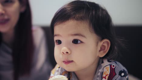 an adorable cute asian toddler boy is eating cookie beside his young mother with red hair.