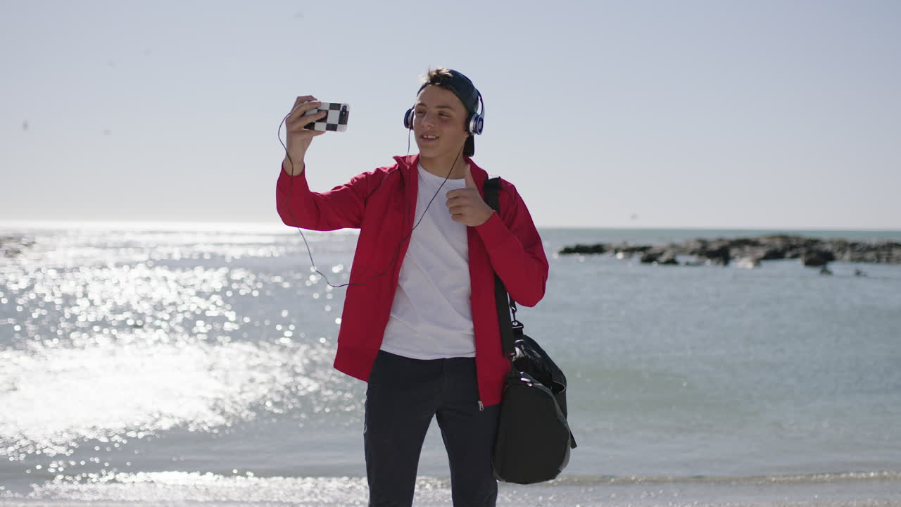un adolescente sonriente toma una foto de selfie usando el teléfono en la playa con una chaqueta roja